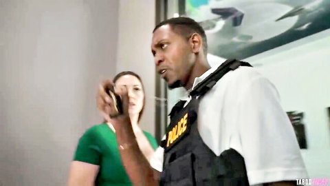Muscular black cop in police vest talks on phone as brunette woman in green top watches intently