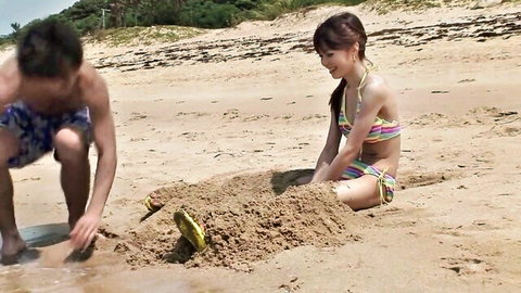 Skinny Japanese teen in colorful bikini building sandcastle on beach with guy
