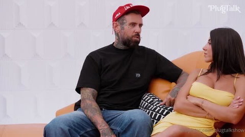Delilah Raige in yellow dress sitting with tattooed man on orange couch
