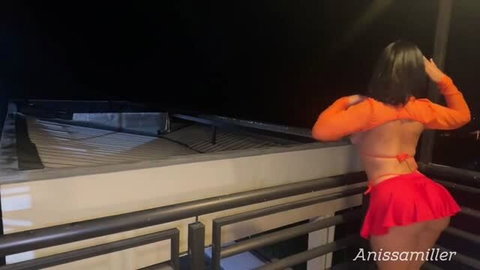 Velma in orange crop top and red skirt posing on balcony at night