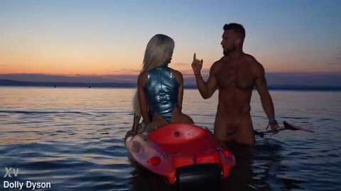 Dolly Dyson in shiny blue latex swimsuit with muscular man in water at sunset