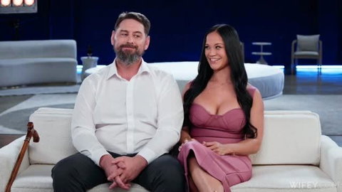 Jazmine Cruz in pink dress sitting with bearded man on white couch