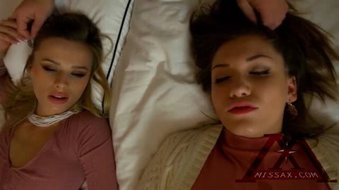 Two attractive women lying side by side on white sheets, one in pink top, one in brown turtleneck