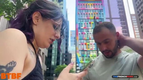Purple haired woman in black top shows phone to bearded man outdoors
